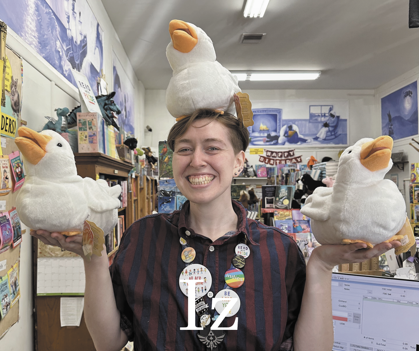 Iz staff photo. A white person with short dirty blonde hair balancing a white duck puppet in each hand and on top of their head wearing a navy and maroon striped button up. "Iz" is written large in the bottom in white.